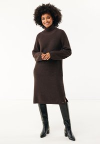 Abito a maniche lunghe a collo alto in marrone scuro con costine e spacchi laterali, abbinato a stivali neri lucidi fino al ginocchio. Tessuto liscio e design aderente.