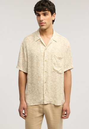 Mann trägt beiges, kurzärmliges Hemd mit dezentem Blumenmuster und beiger Hose, steht vor einfarbigem Hintergrund.