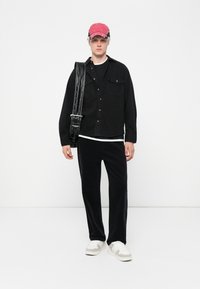 Model trägt ein schwarzes Denim-Hemd, schwarze Cordhosen und eine rote Mütze. Außerdem hat sie eine schwarze Umhängetasche mit silbernen Akzenten und weiße Sneakers.