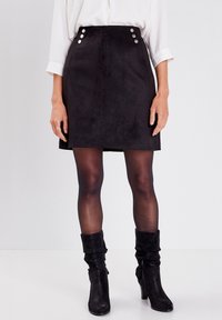 Zwarte suède mini-jurk met een aansluitende silhouet, voorzien van vijf zilveren knopen op de hoge tailleband. Draagt het met doorzichtige zwarte panty’s en enkellaarsjes.