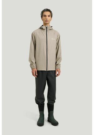 Mand iført beige hætte regnjakke, sorte vandtætte bukser og grønne gummistøvler, stående mod en ensfarvet lys baggrund.