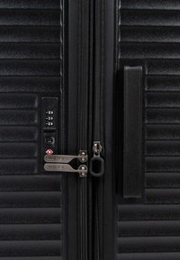 Valigia rigida nera con righe orizzontali, dotata di due cerniere argentate, una serratura a combinazione e una superficie testurizzata.