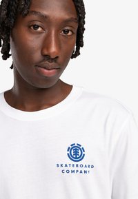 Camiseta de algodón blanca con un logo gráfico azul que lleva el texto "SKATEBOARD COMPANY" en el área del pecho izquierdo. Cuello redondo y mangas cortas.