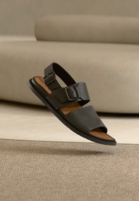 Μαύρα δερμάτινα σανδάλια με δύο ρυθμιζόμενες πόρπες, ανοιχτή μύτη και καφέ επενδεδυμένο πάτο, τοποθετημένα πάνω σε ουδέτερο φόντο.