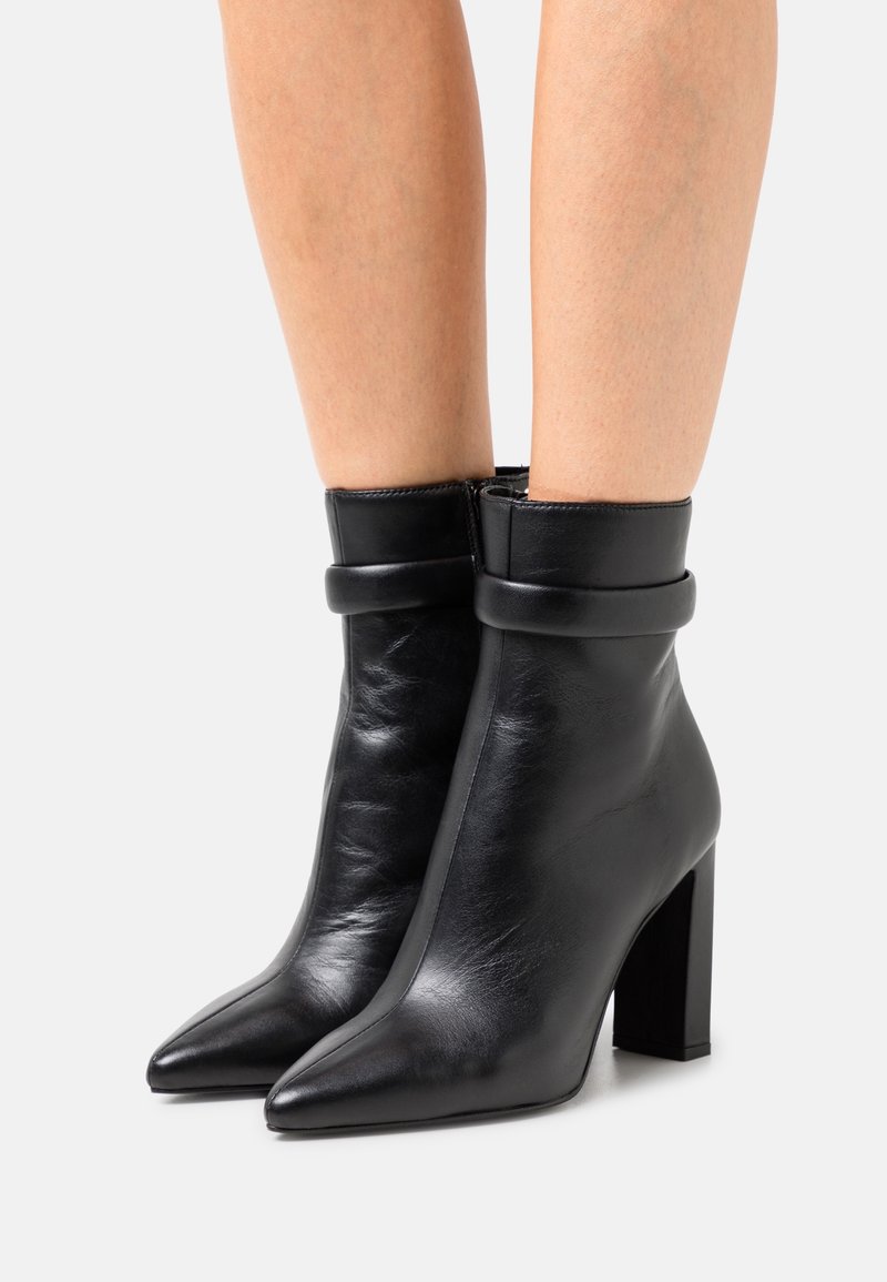 Jambes portant des bottines en cuir noir avec des revers pliés, des bouts pointus et des talons carrés sur fond blanc.