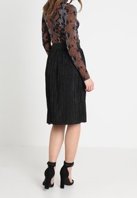 Svart plisserad kjol med en strukturerad yta, som har ett brett midjeband. Modellen har på sig svarta ankelsandaler med remmar, vilket visar hud ovanför knäet.