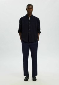 Granatowa koszula zapinana na guziki z podwiniętymi rękawami, zestawiona z ciemnymi spodniami i czarnymi butami. Gładki materiał, dopasowany krój, minimalistyczny design.