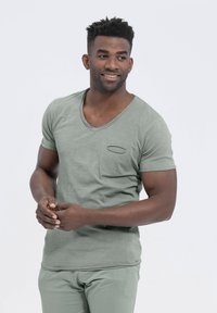 Camiseta verde de cuello en V con mangas cortas y un pequeño bolsillo en el pecho. La tela parece suave con un acabado ligeramente texturizado y un diseño ajustado.