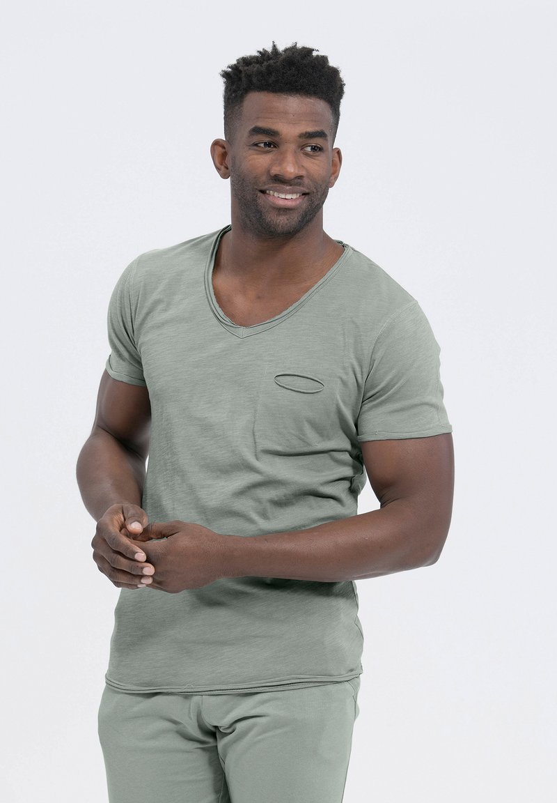 Camiseta verde de cuello en V con mangas cortas y un pequeño bolsillo en el pecho. La tela parece suave con un acabado ligeramente texturizado y un diseño ajustado.