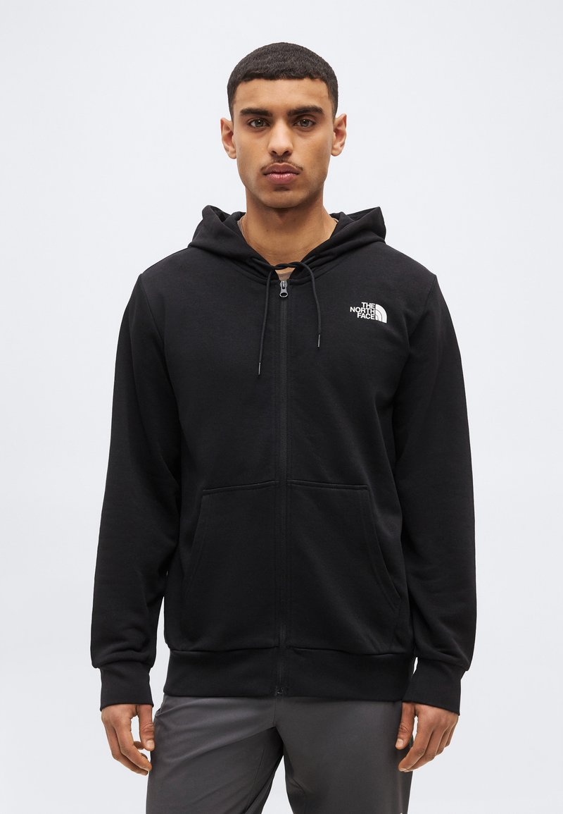 Νεαρός άνδρας με κοντά μαλλιά, φορώντας μαύρο φούτερ με φερμουάρ και το λογότυπο The North Face, στέκεται μπροστά σε απλό φωτεινό φόντο.