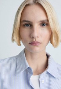 Loose-fitting, collared shirt with light blue and white vertical stripes, made of a smooth fabric, featuring a buttoned placket and subtle texture.