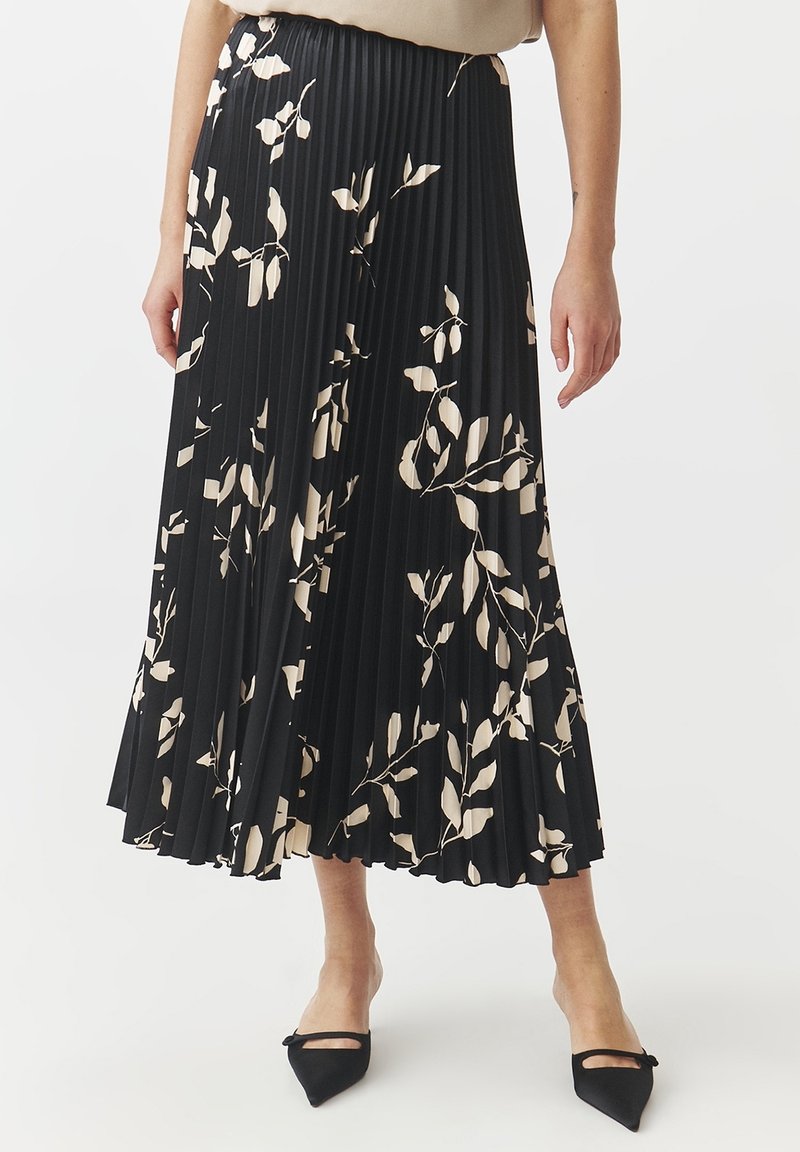 Jupe midi plissée noire avec motifs de feuilles beiges portée avec un top beige et des ballerines noires à bouts pointus avec des brides.