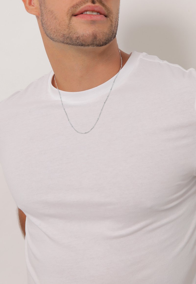 Tricou alb cu mânecă scurtă, realizat dintr-un material moale și neted. Are un colier subțire din lanț argintiu, cu detalii de mici mărgele.