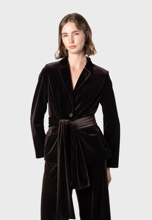 Jeune femme portant un blazer en velours noir avec une longue ceinture nouée à la taille et un pantalon noir assorti sur un fond uni.