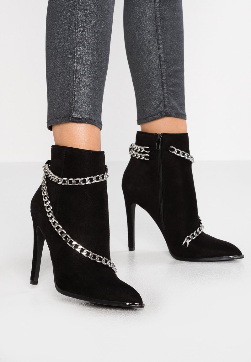 Bottines en daim noir à talons hauts avec des détails en chaîne argentée et une fermeture éclair sur les côtés intérieurs, portées avec un pantalon ajusté gris foncé scintillant.