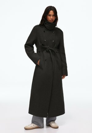 Femme portant un long manteau sombre à double boutonnage avec ceinture nouée à la taille, debout avec une main dans la poche, portant un jean gris et des baskets blanches.