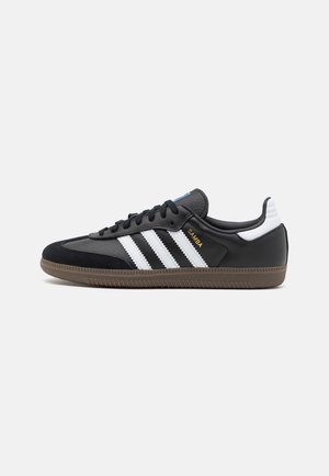 Baskets noires Adidas Samba avec des bandes blanches, semelle en gomme, lacets noirs et texte doré "Samba" sur le côté, montrées de la face extérieure.