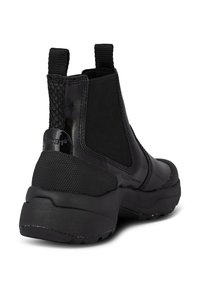 Botte Chelsea noire épaisse avec talon texturé, panneaux latéraux élastiques et languettes de traction sur fond blanc.