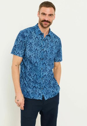 Mann mit Schnurrbart, der ein blau gemustertes kurzärmeliges Hemd und dunkle Hose trägt, steht mit einer Hand in der Tasche.