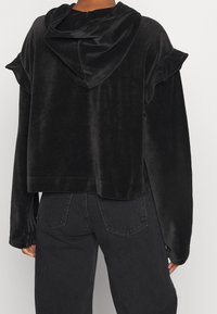 Kort svart sammetströja med en mjuk textur, vågiga axelsömmar och en draperad huva. Matchad med högmidjade svarta jeans.