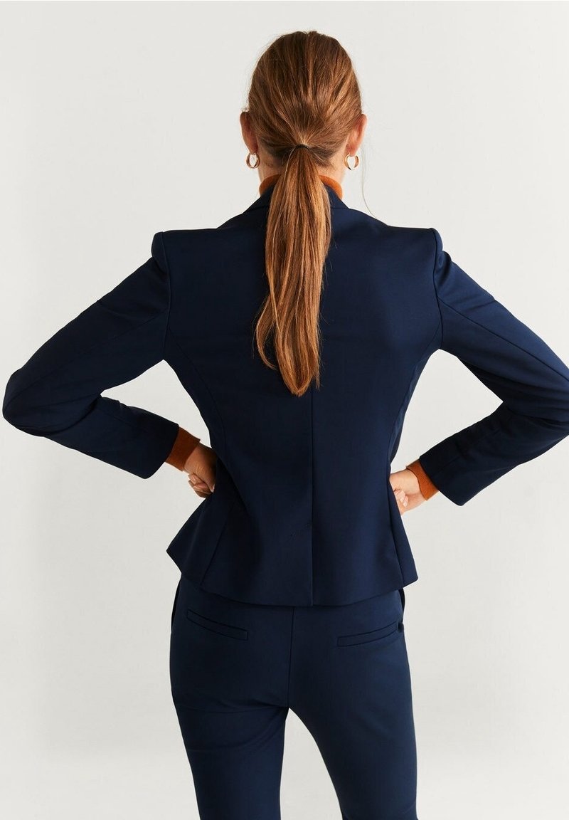 Femme aux longs cheveux bruns en queue de cheval, portant un costume bleu marine, debout les mains sur les hanches, de dos à la caméra.