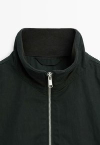 Black jacket featuring a high collar, front zipper closure, and smooth fabric texture. Stitching detail along collar and zipper.
