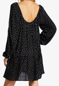 Žena v černých šatech s bílými puntíky, dlouhými nafouklými rukávy a hlubokým výstřihem na zádech, který odhaluje horní část zad.