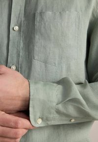 Jasnozielona lniana koszula z kołnierzykiem zapinanym na guziki, pojedynczą kieszenią na piersi i mankietami zapinanymi na guziki. Miękka textura z widocznym splotem materiału.