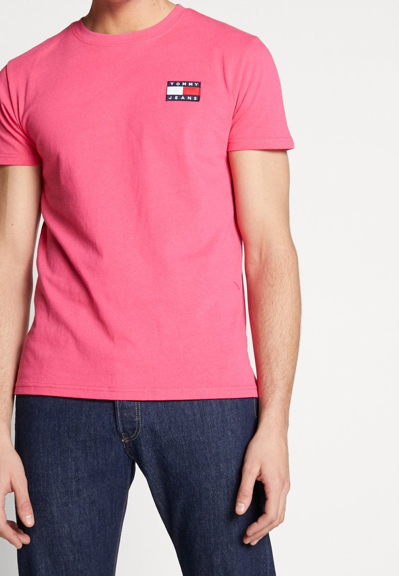 Pink cotton T-shirt featuring a small blue, white, and red embroidered logo patch on the left chest; short sleeves and crew neck design.