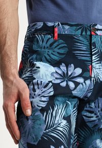 Blomstrede shorts i mørk navy, med livlige grønne og lyseblå bladmønstre. Indeholder røde accenter på lommerne og knaplukning.