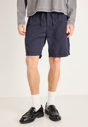 Pantalones cortos azul marino de tela ligera con cintura ajustable con cordón. Combinados con mocasines negros y calcetines blancos hasta el tobillo.