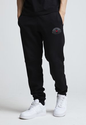 Osoba ubrana w czarne spodnie dresowe z logo Zavetti na udzie i białe trampki, stojąca z rękami w kieszeniach na jednolitym tle.