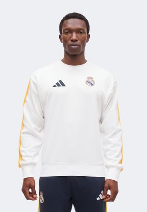 Homme portant un sweat-shirt blanc du Real Madrid avec des rayures jaunes sur les manches et un pantalon bleu marine, debout devant un fond uni.