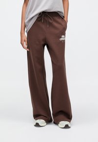 Καφέ φαρδιά φόρμα με ελαστική μέση και κορδόνι ρύθμισης. Φέρει το λογότυπο «Nike Athletics» σε λευκό. Κατασκευασμένη από μαλακό, υφασμάτινο ύφασμα με υφή.