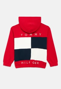 Κόκκινο φούτερ με κουκούλα με σχέδιο σε λευκό και ναυτικό καρό στην πλάτη. Φέρει το λογότυπο "TOMMY HILFIGER" με λευκά γράμματα· μαλακό ύφασμα και ελαστικές μανσέτες.