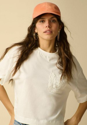 Jeune femme aux longs cheveux ondulés porte une casquette orange, des boucles d'oreilles anneaux et chaîne en or, un t-shirt blanc avec une poche crochetée.
