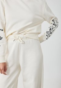 Sudadera color crema con bordado floral negro en las mangas, combinada con pantalones de cordón a juego que presentan un ajuste relajado y una cintura elástica.