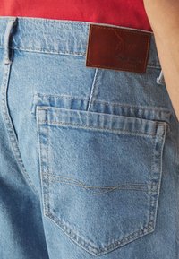 Heledad sinised teksad, millel on pruun nahklabel vööl ja kaks tagumist taskut nähtavate õmbluste detailidega.