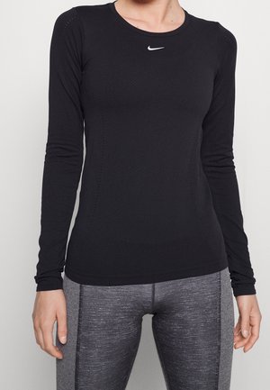 Person trägt ein schwarzes Nike-Oberteil mit langen Ärmeln und graue, sportliche Leggings und steht vor einem einfachen, hellen Hintergrund.