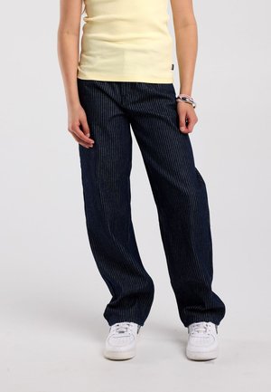 Person wearing dark pinstripe wide-leg pants, white sneakers, a light yellow sleeveless top, and bracelets on the right wrist.