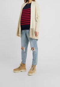 Beige Cardigan mit Fransen, marineblauer gestreifter Pullover mit roten Linien, ausgebleichte hellblaue Jeans und beige Schnürstiefel mit dicken Sohlen.