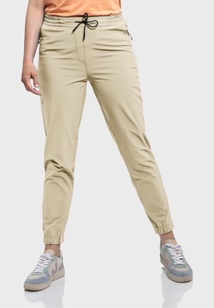 Personne portant un pantalon jogger beige avec des poignets élastiques et un cordon noir, associé à des baskets bleues et beiges sur un fond clair.