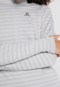 Světlounce šedý pulovr na zip s vodorovnými pruhy, texturovanou tkaninou a logem Salomon na hrudi. Má dlouhé rukávy a vysoký límec.