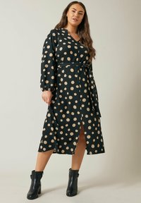 Schwarzes Kleid mit goldenen Punkten, V-Ausschnitt, langen Ärmeln und tailliertem Bund. Verfügt über einen vorderen Schlitz und einen strukturierten Stoff. Getragen mit schwarzen Ankle-Boots.