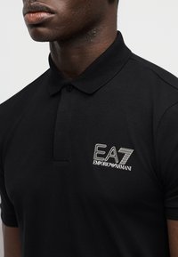 Μαύρο πόλο μπλουζάκι από μαλακό ύφασμα με κλασικό γιακά και λευκό κεντημένο λογότυπο "EA7 EMPORIO ARMANI" στο αριστερό στήθος.