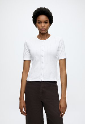 Femme aux cheveux courts et bouclés portant un haut blanc à manches courtes boutonné et un pantalon taille haute foncé sur un fond uni.