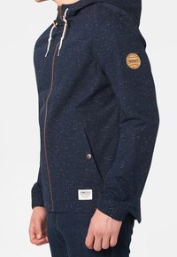 Marinblå kapuschongjacka med spräcklig textur, framfickslås, dragsko i huvan och en logotyp-patch på ärmen. Har sidofickor.