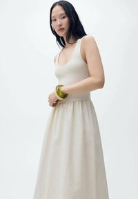 Robe sans manches beige avec un corsage ajusté et une jupe évasée, fabriquée à partir d'un tissu lisse ; associée à un bracelet translucide vert.