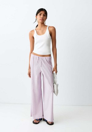 Vrouw draagt een witte mouwloze top en lichtpaarse wijde broek, houdt een grijze gestructureerde clutch vast, staand tegen een witte achtergrond.
