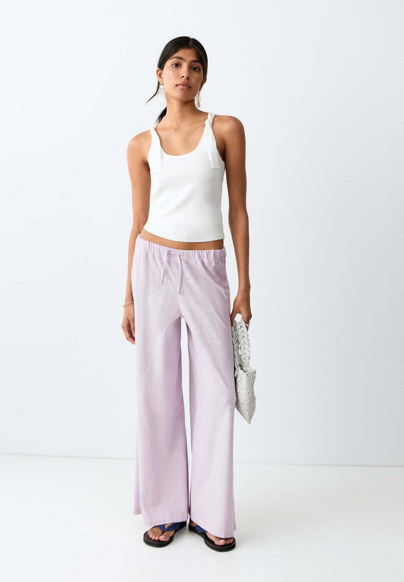 Femme portant un haut sans manches blanc et un pantalon large violet clair, tenant une pochette grise texturée, debout sur fond blanc.